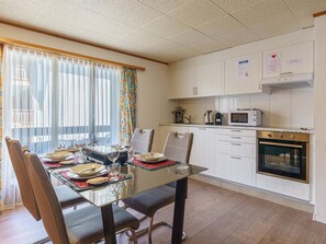 A dining area with a glass table and chairs, a kitchen with white cabinets and appliances, and a window with sheer curtains. - Haus Slalom Appartement/Fewo Apt #4 by Interhome (Saas-Fee)