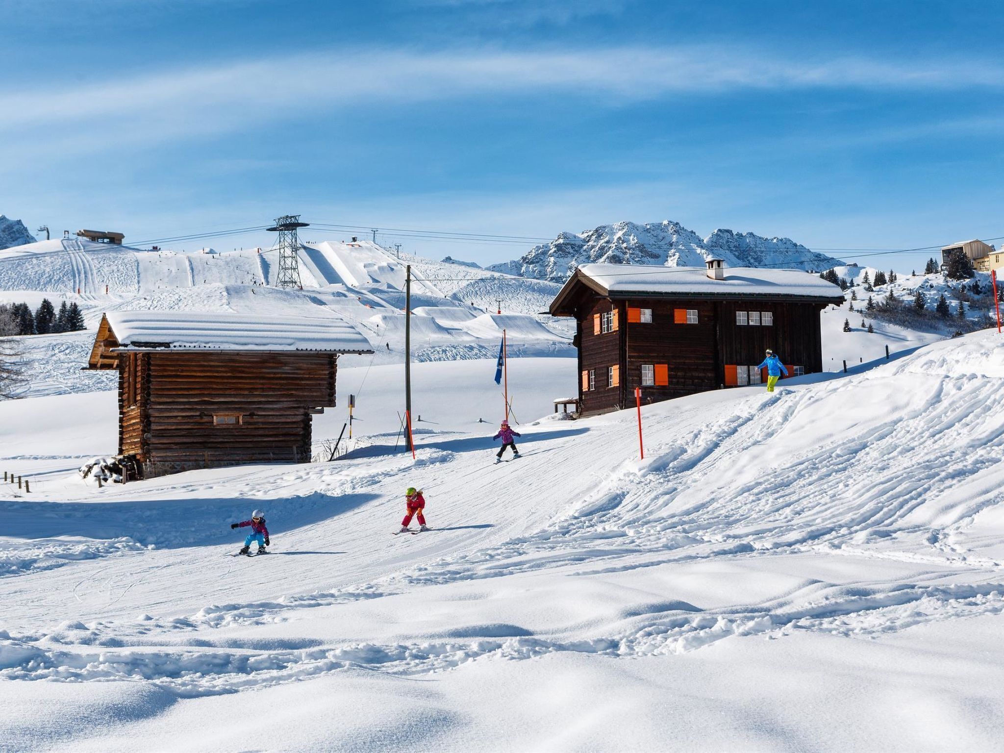 A wooden cabin with a snow-covered roof, a ski slope with skiers, and a mountain backdrop.
