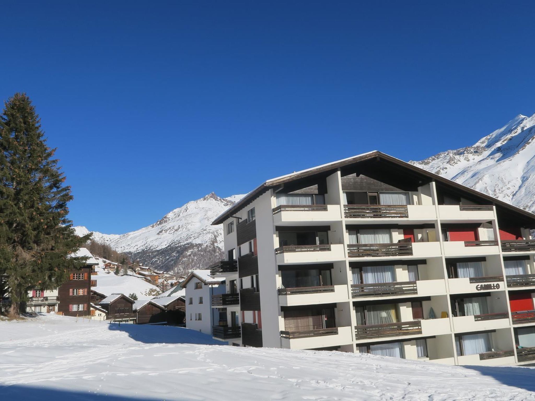 Ein mehrstöckiges Hotel mit Balkonen, im Hintergrund eine Skipiste und schneebedeckte Berge.