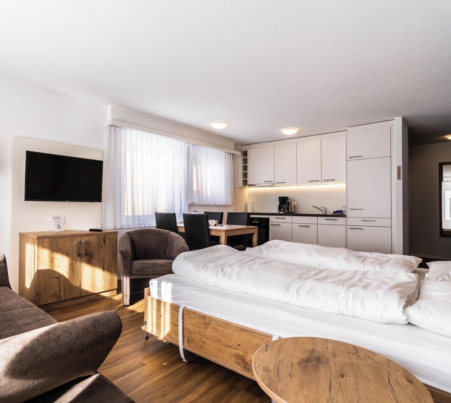 A modern hotel room with a bed, a desk, a chair, a television, and a kitchenette.