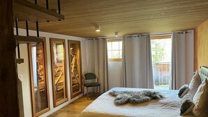 Ein Schlafzimmer mit Bett, Sessel, Holzboden und einem Fenster mit Vorhängen.