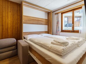 A bedroom with a wooden headboard, a bed with white linens, a bench, and a window with curtains. - Haus Slalom Appartement/Fewo Apt #7 by Interhome (Saas-Fee)