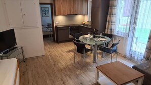 Uma sala de estar moderna com uma mesa de jantar de vidro, cadeiras pretas, uma mesa de centro de madeira e uma TV sobre um suporte.