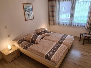 Ein Schlafzimmer mit einem Bett, einem Nachttisch mit Lampe, einem Sessel und einem Fenster mit Vorhängen.