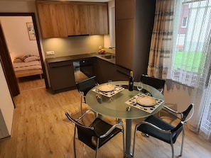 Un espace de vie compact comprenant un coin repas avec une table en verre et des chaises noires, une kitchenette dotée de meubles en bois et un lit à l’arrière-plan.