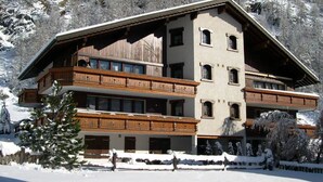 Um edifício com várias andares e varandas, cercado por árvores e montanhas cobertas de neve.