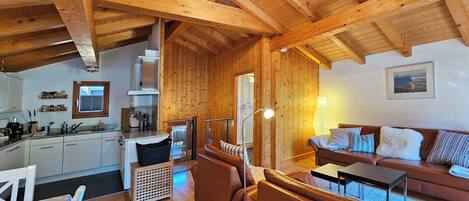 A spacious living area with a wooden ceiling, a kitchen with white cabinets, a sofa with patterned cushions, and a dining table with chairs.