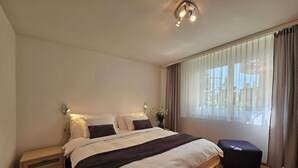 Een slaapkamer met een groot bed, nachtkastjes, een raam met gordijnen en een plafondlamp.