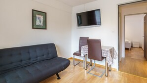 Ein modernes Wohnzimmer mit einem schwarzen Sofa, einem Esstisch mit zwei Stühlen, einem flachbildfernseher an der Wand und einem gerahmten Bild.