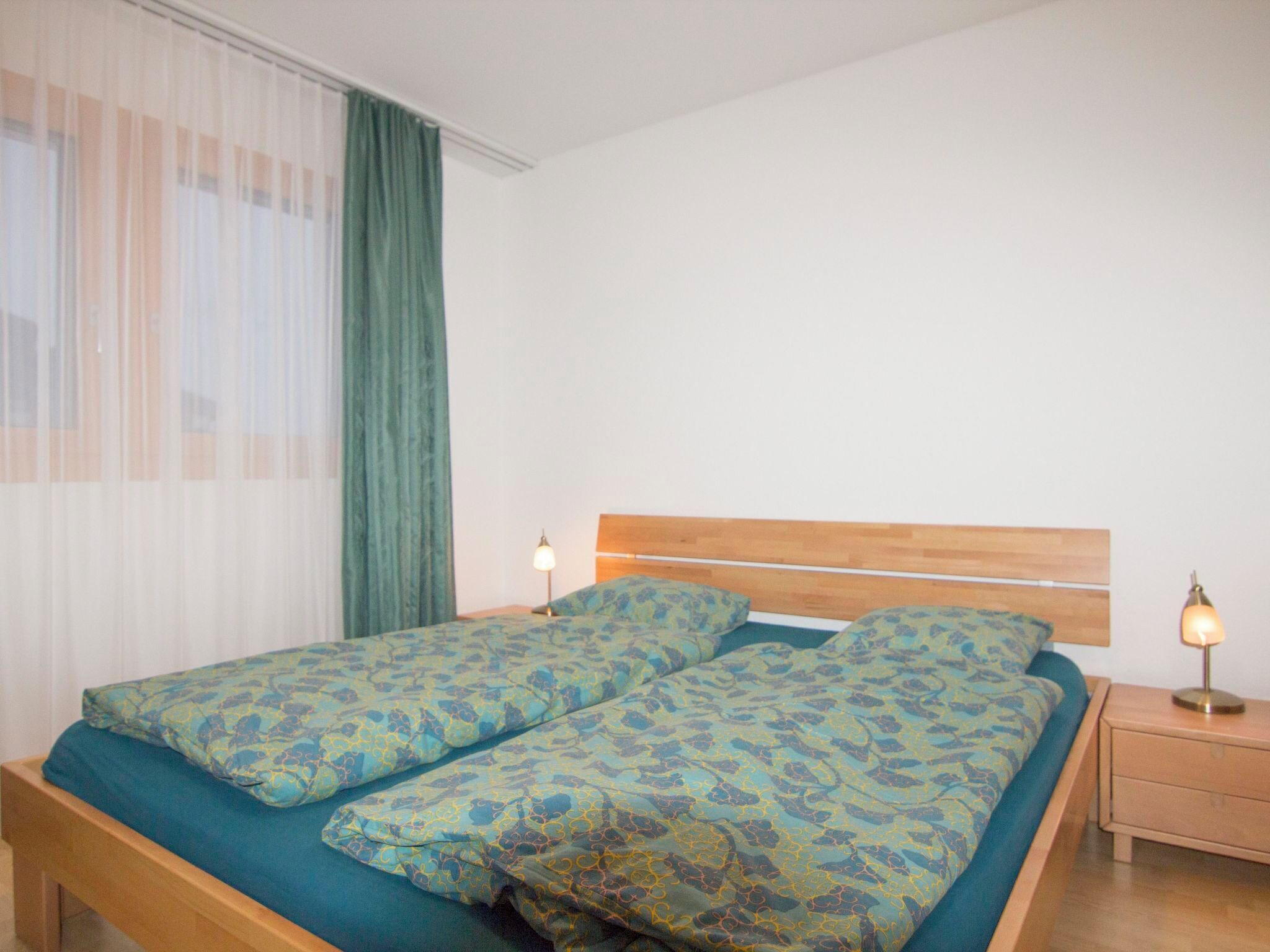 A double bed with patterned bedding, a wooden headboard, two bedside lamps, and a window with sheer curtains.