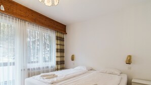 A hotel room with a bed, two bedside tables, a wooden ceiling, and a window with sheer curtains. - Haus City Appartement/Fewo Apt #6 by Interhome (Saas-Grund)