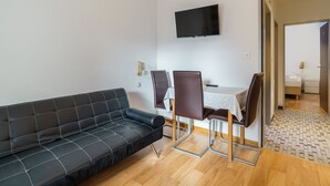 A modern living room with a black sofa, a dining table with chairs, a flat-screen TV mounted on the wall, and a visible door leading to a bedroom. - Haus City Appartement/Fewo Apt #6 by Interhome (Saas-Grund)