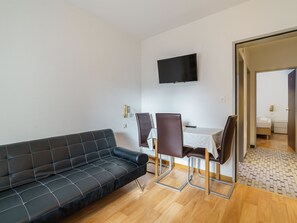 Ein modernes Wohnzimmer mit einer schwarzen Couch, einem Esstisch mit Stühlen, einem flachbildfernseher an der Wand und einer sichtbaren Tür zu einem Schlafzimmer.