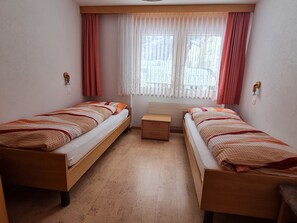 Ein Zimmer mit zwei Holzbetten, einem kleinen Nachttisch, einem Fenster mit Vorhängen und einer Wandleuchte.