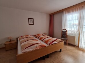 A bedroom with a wooden bed, a nightstand, a chair, and a window with curtains. - Orion Appartement/Fewo 4-Bettwohnung Nr. 53 by Interhome (Saas-Grund)