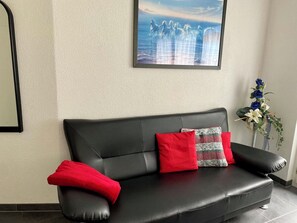 Ein schwarzes Ledersofa mit roten und grauen Kissen, ein gerahmtes Kunstwerk darüber und ein Blumentopf mit blauen und weißen Blüten zur Rechten.