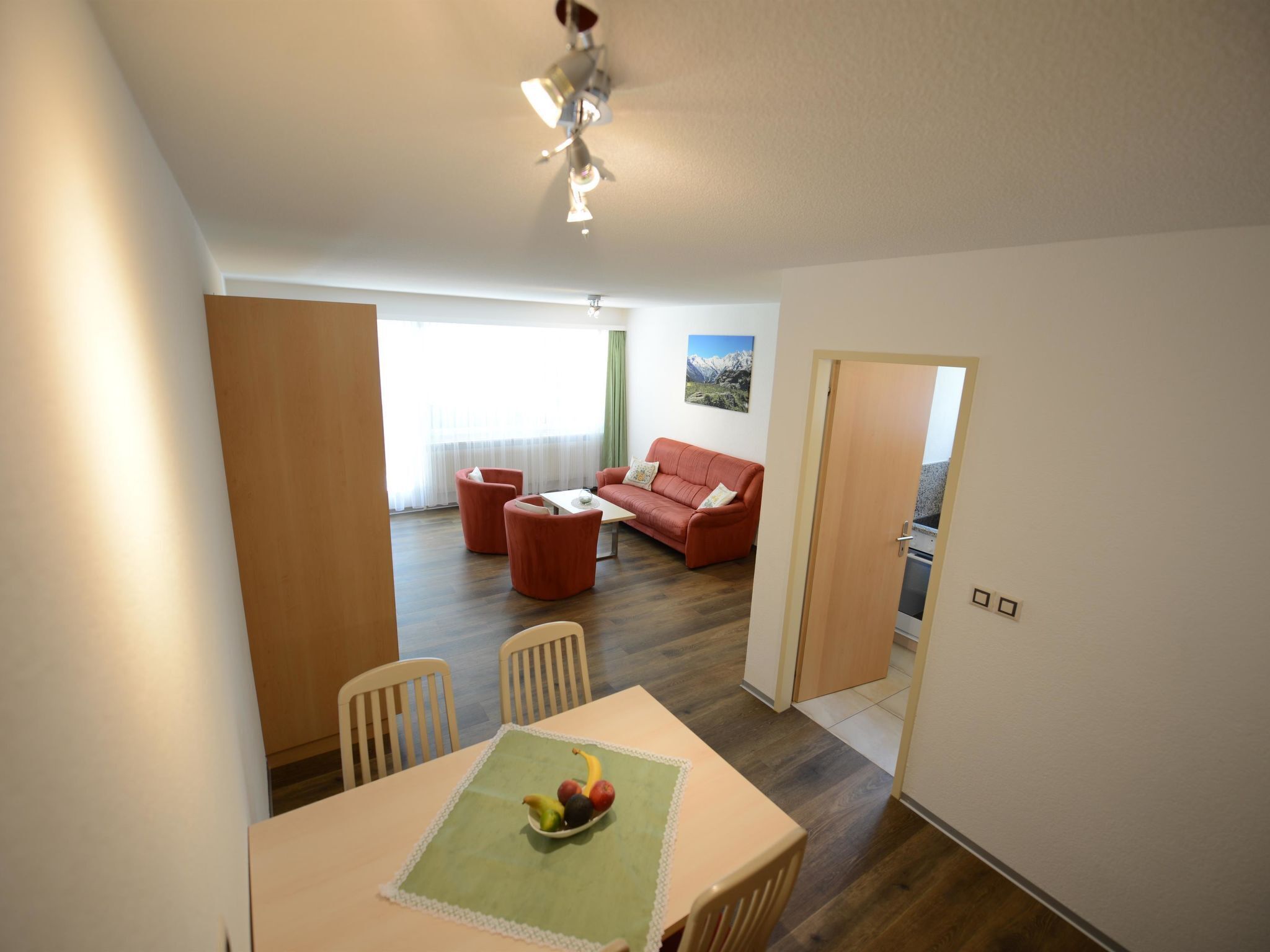 Ein Essbereich mit einem Tisch für zwei Personen, eine rote Couch im Hintergrund und Blick auf eine Berglandschaft durch das Fenster.