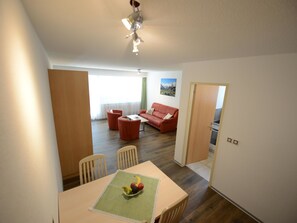 Ein Essbereich mit einem Tisch für zwei Personen, eine rote Couch im Hintergrund und Blick auf eine Berglandschaft durch das Fenster.
