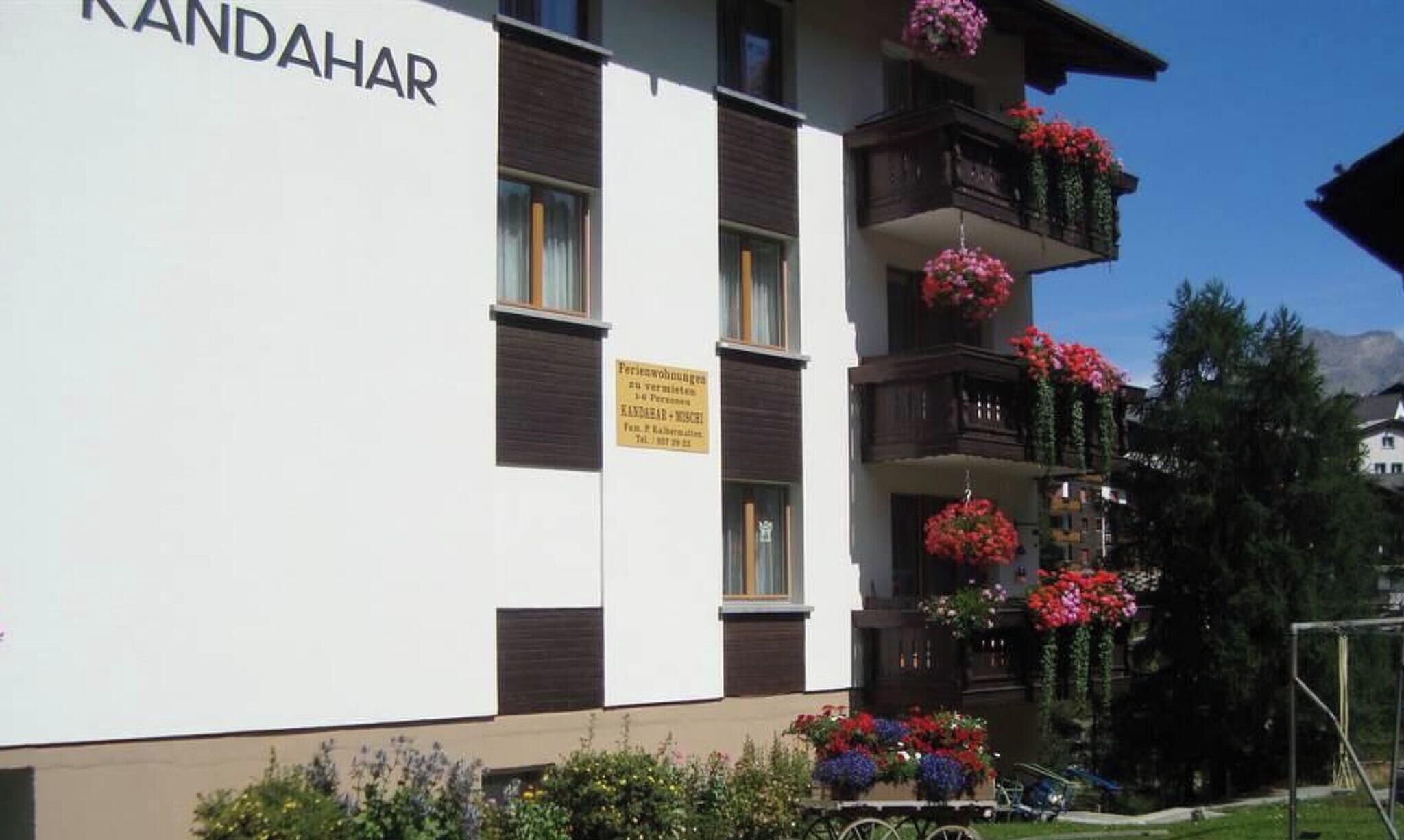 Ein mehrstöckiges Gebäude mit Balkonen, die mit Blumen geschmückt sind, ein Schild, das auf eine Ferienwohnung hinweist, und ein klarer Himmel im Hintergrund.