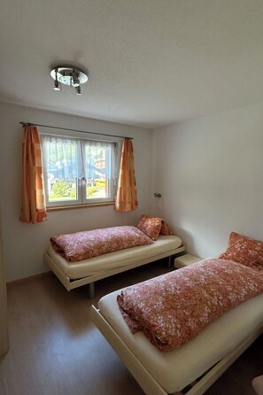 A small, single-room accommodation with two beds, a window with curtains, and a ceiling light fixture. - Alpenstern Appartement/Fewo 4-Bettwohnung "Distel" by Interhome (Saas-Grund)