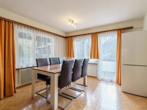 A modern dining area with a wooden table and chairs, large windows with curtains, and a white refrigerator. - Haus City Appartement/Fewo Apt #3 by Interhome (Saas-Grund)