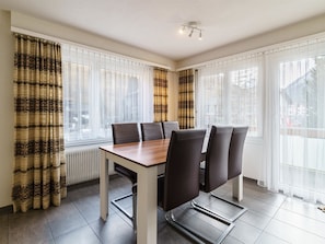 A dining area with a table and chairs, large windows with curtains, and a radiator. - Haus City Appartement/Fewo Apt #1 by Interhome (Saas-Grund)