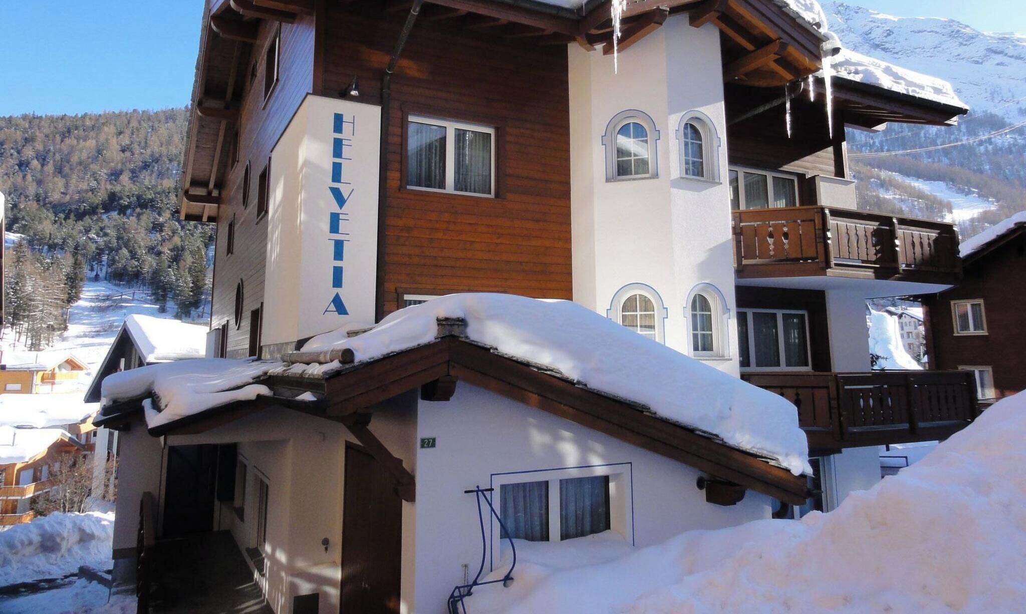 Un hôtel à plusieurs étages, avec une enseigne portant l’inscription « HEVETIA » sur la façade, des balcons en bois et un paysage enneigé en arrière-plan.