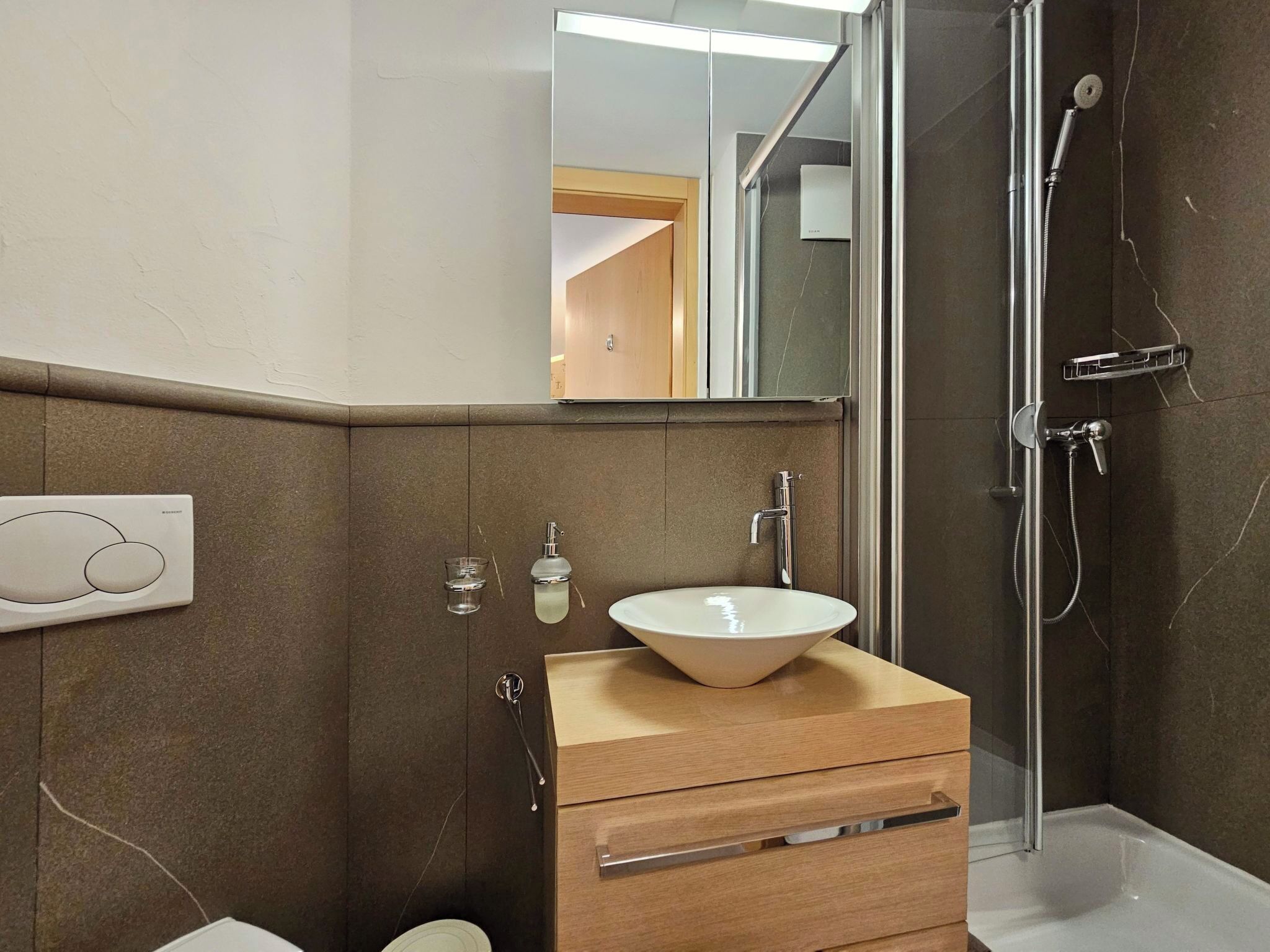 Un baño moderno con ducha compartimentada por vidrio, tocador de madera con lavamanos blanco y espejo sobre el tocador.