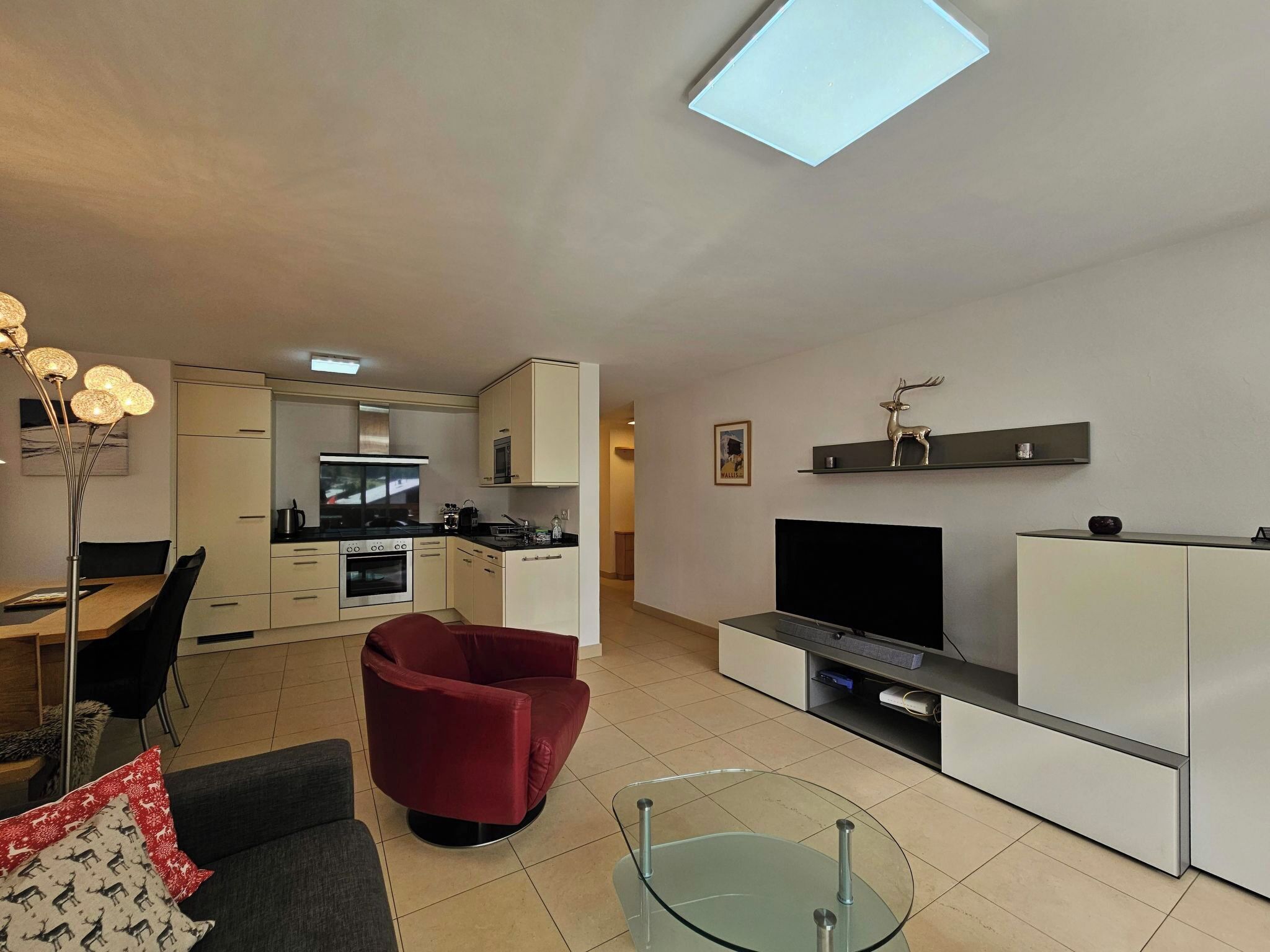Una sala de estar moderna con una mesa de centro de vidrio, un sillón rojo, un televisor y una cocina al fondo.