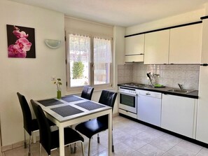 Cocina compacta con armarios blancos, estufa y lavamanos. Hay una mesa de comedor con sillas y un florero con flores en la pared.