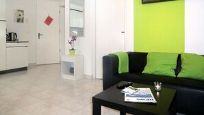 Ein Wohnzimmer mit einer schwarzen Couch, einem kleinen Tisch mit Fernbedienung und einer Zeitschrift. Im Hintergrund ist eine grüne Wand mit einem Bilderrahmen und eine weiße Tür zu sehen.