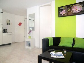 Un salón con un sofá negro, una mesita con un control remoto y una revista. Se aprecia una pared verde con un marco de fotos y una puerta blanca al fondo.