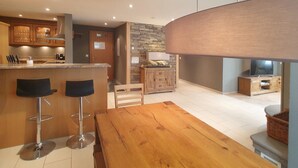 Une cuisine moderne avec un mur d’accent en pierre, des tabourets de bar en bois, une table à manger en bois et un téléviseur placé dans un meuble en bois.