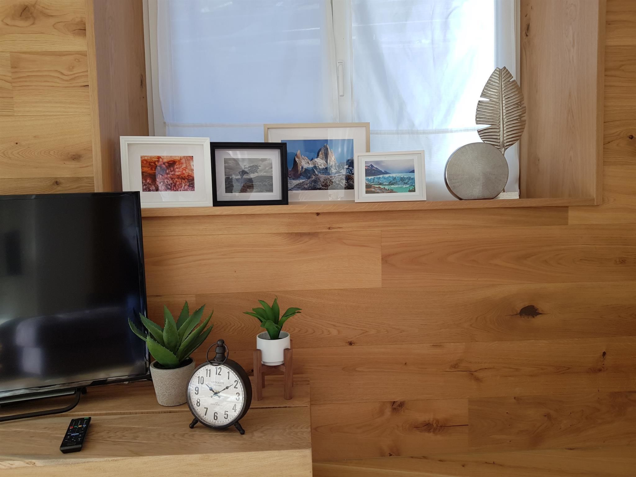 Un intérieur en bois avec une télévision, deux plantes en pot, une horloge et des photos encadrées disposées sur une étagère.