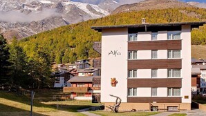 Ein modernes Gebäude mit der Aufschrift 'Alfa' auf der Fassade, umgeben von einer gebirgigen Landschaft mit schneebedeckten Gipfeln und einem bewaldeten Gebiet.