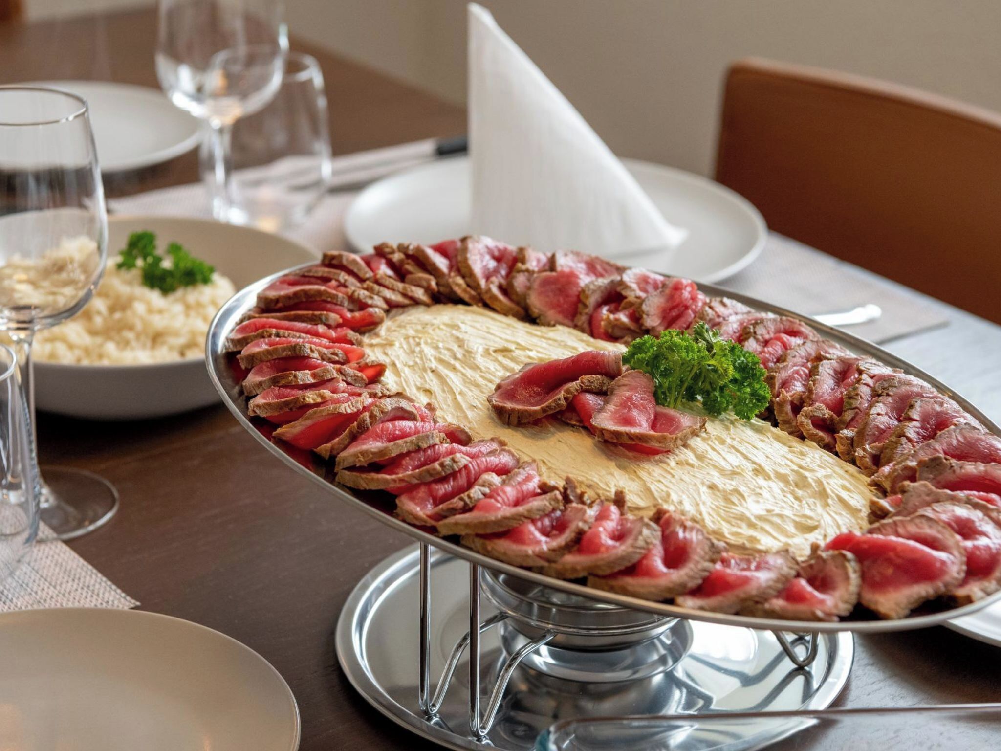 A round metal serving tray with sliced meat arranged in a circular pattern, garnished with parsley, on a table with a white napkin and a glass of water.