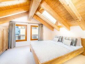 A bedroom with a sloped ceiling, wooden beams, a bed with pillows, and two windows with curtains. - Sonnegg Penthouse Appartement/Fewo 10-Bettwohnung by Interhome (Saas-Fee)