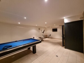 Ein Billardtisch mit Spielstöcken, eine Uhr und ein Fernseher an der Wand in einem Raum mit Fliesenboden und eingelassener Beleuchtung.
