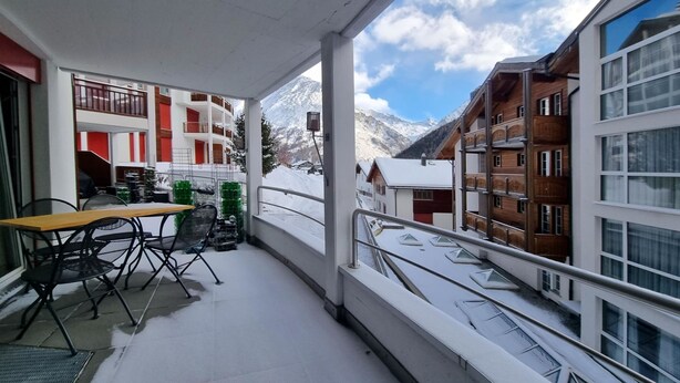 Un balcon avec une table et des chaises, une balustrade, et une vue sur des montagnes et des bâtiments enneigés.