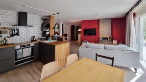 Une cuisine moderne avec des carreaux blancs, une table à manger en bois et un coin salon agrémenté d’un mur d’accent rouge et d’une cheminée.