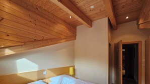 Un dormitorio con techo de madera, una cama con ropa blanca, una lámpara de noche y una puerta que da a otra habitación.