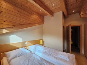 A bedroom with a wooden ceiling, a bed with white bedding, a bedside lamp, and a door leading to another room. - Aramis 4. OG Appartement/Fewo Aramis 4. OG | 6-Bettwohnung by Interhome (Saas-Fee)