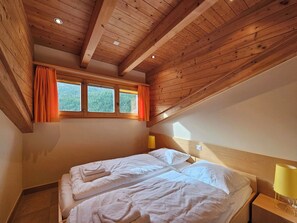 A bedroom with a wooden ceiling, a bed with white linens, and a window with orange curtains. - Aramis 4. OG Appartement/Fewo Aramis 4. OG | 6-Bettwohnung by Interhome (Saas-Fee)