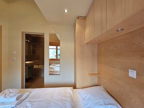 A bedroom with a wooden headboard, a bed with white linens, a small bedside table, and a mirror reflecting a bathroom with a shower. - Aramis 4. OG Appartement/Fewo Aramis 4. OG | 6-Bettwohnung by Interhome (Saas-Fee)