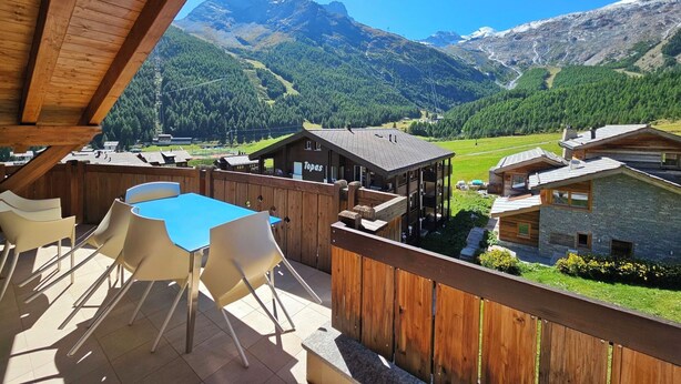 Una terraza de madera con mobiliario exterior, vistas a la montaña y un edificio con la leyenda 'Topas'.