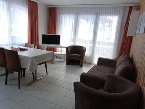 Una sala de estar moderna con comedor, televisor de pantalla plana y ventanas amplias con cortinas.