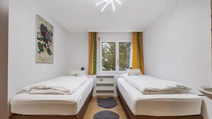 Ein Zimmer mit zwei Betten, einem Fenster mit Vorhängen, einem kleinen Tisch und einem Wandbild.