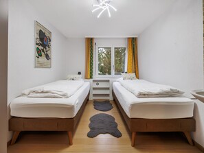 A room with two beds, a window with curtains, a small table, and a wall-mounted artwork. - Everest Appartement/Fewo 4-Bettwohnung K by Interhome (Saas-Fee)