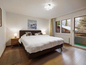 Ein Schlafzimmer mit einem großen Bett, einem Nachttisch mit Lampe, einem Bild an der Wand und einem Fenster mit Blick auf Bäume.