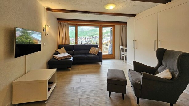 Ein Wohnzimmer mit Sofa, Sessel, Fernseher und Blick auf eine Stadt durch ein großes Fenster.